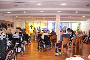 Versammlung im Gemeindezentrum mit Senioren in Rollstühlen und Stühlen sowie einer Ansprache vorne.