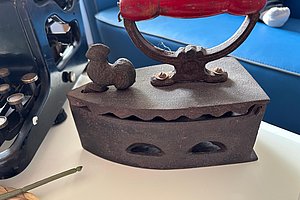 Altes Bügeleisen aus Metall mit rotem Holzgriff auf weißer Oberfläche neben einer schwarzen Schreibmaschine