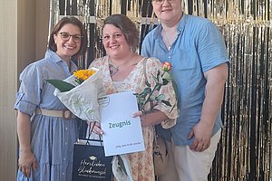 Drei Personen vor gold-weißer Lamettadekoration, eine Person hält Blumenstrauß und Dokument mit der Aufschrift 'Zeugnis'