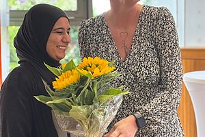 Zwei Frauen stehen nebeneinander, eine hält einen Strauß Sonnenblumen, die andere trägt ein schwarz-weiß gemustertes Kleid mit V-Ausschnitt und eine Armbanduhr.