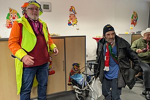 Zwei Männer in bunten Karnevalskostümen, einer mit Rollator, in fröhlicher Feier-Atmosphäre.