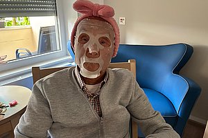 Person mit grauem Pullover und rosa Haarband sitzt an einem Tisch in einem Raum mit blauem Sessel und Fenster