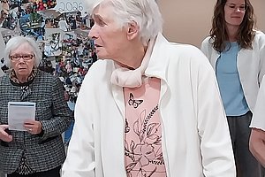 Drei ältere Personen in einem Raum, eine Frau mit weißem Haar und weißer Jacke steht im Vordergrund, eine weitere Frau hält ein Blatt Papier, im Hintergrund eine Pinnwand mit Fotos.