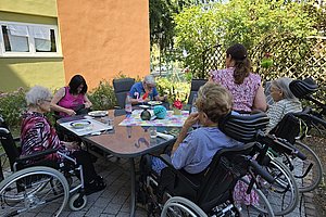 Mehrere ältere Menschen, einige im Rollstuhl, sitzen an einem Gartentisch im Freien und beschäftigen sich mit Bastelmaterialien