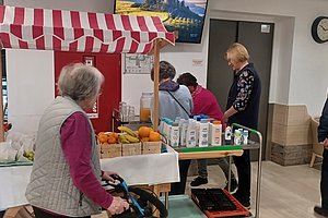 Marktstand mit frischen Früchten und Getränken, Senioren beim Einkaufen in einem Gemeinschaftsraum.