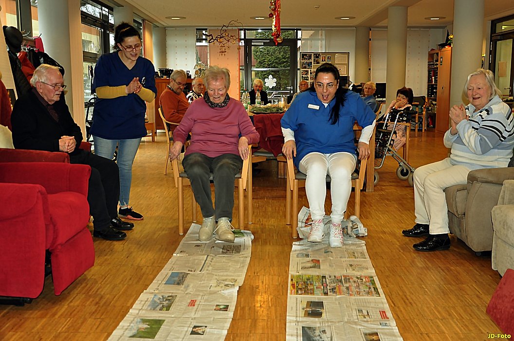 Mehrere ältere Menschen sitzen und stehen in einem Gemeinschaftsraum, zwei von ihnen sitzen auf Stühlen mit Zeitungen auf dem Boden davor, umgeben von weiteren sitzenden und stehenden Personen.