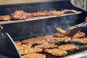 Mehrere gewürzte Fleischstücke liegen auf einem Grillrost und werden mit einer Grillzange gewendet