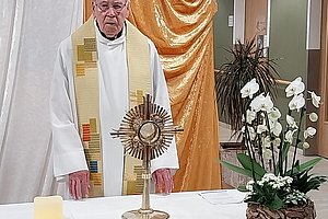 Priester in weißem Gewand mit goldbesticktem Stola steht hinter einem Altar mit Monstranz und Blumengesteck