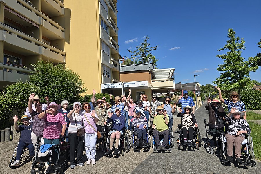 Gruppe älterer Menschen im Freien, fröhlich winkend vor einem Wohnheim unter blauem Himmel.