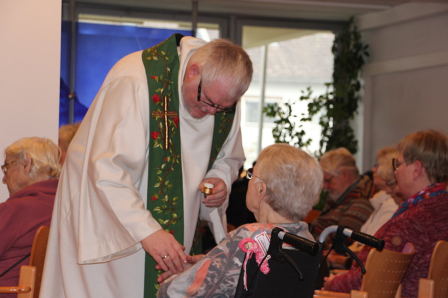 Ein Priester in weißem Gewand mit grünem Stola reicht einer sitzenden älteren Frau in Rollstuhl eine Hostie in einer Kapelle oder einem Gemeinschaftsraum.