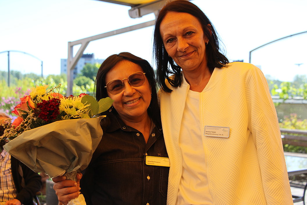 Zwei Frauen mit Namensschildern, eine hält einen großen Blumenstrauß, beide stehen in einem hellen Raum mit Fenster und Blick ins Grüne.