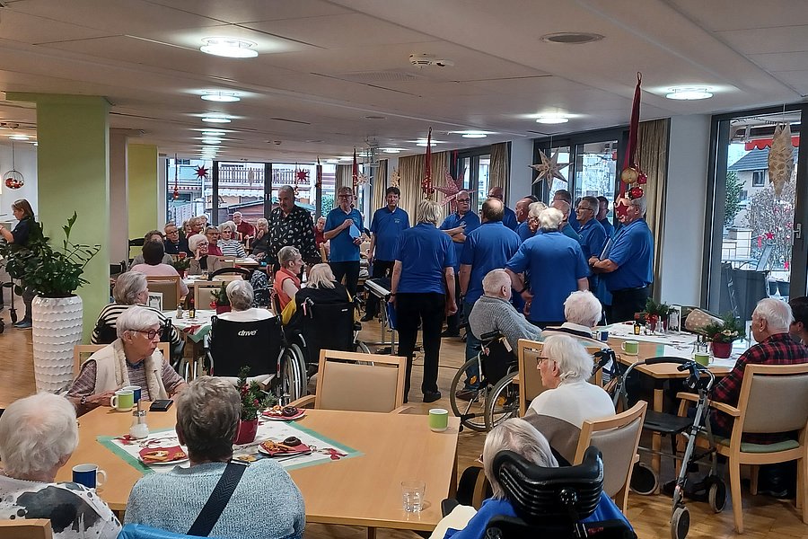 Gemütliche Atmosphäre in einem Gemeinschaftsraum mit Gästen und einem Männerchor in blauen Shirts.