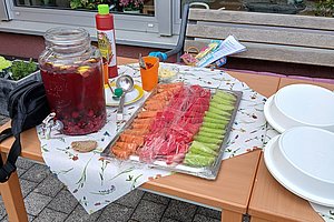 Tisch mit Karaffe gefüllt mit rotem Getränk, Plastikbechern, Tablett mit geschnittenem Obst und mehreren weißen Tellern auf einer Terrasse vor einem Fenster