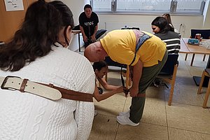 Mehrere Personen sitzen in einem Raum, eine Person in gelbem T-Shirt beugt sich nach vorne, eine andere hält deren Fuß.