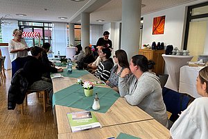 Mehrere Personen sitzen an einem langen Tisch mit grünen Tischläufern in einem hellen Raum und hören einer stehenden Frau zu