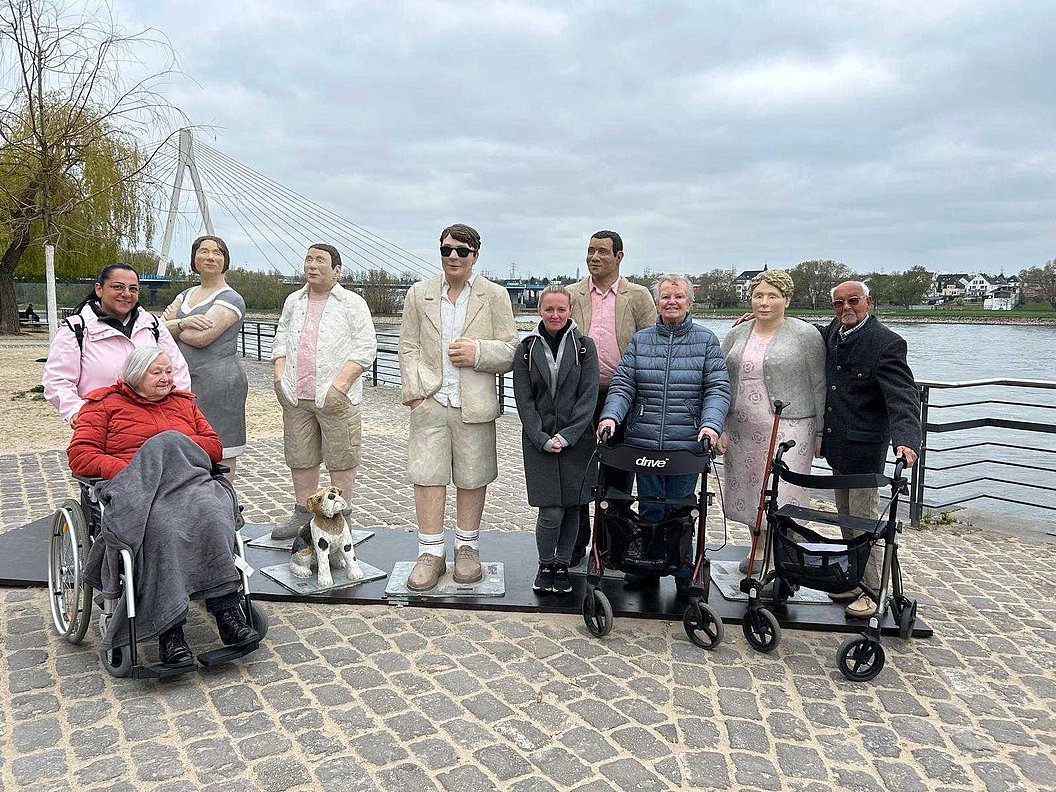 Gruppe von Menschen, darunter zwei mit Rollstuhl, zwei mit Rollator, und mehrere stehende Personen auf gepflastertem Platz an einem Flussufer mit einer Hängebrücke im Hintergrund.