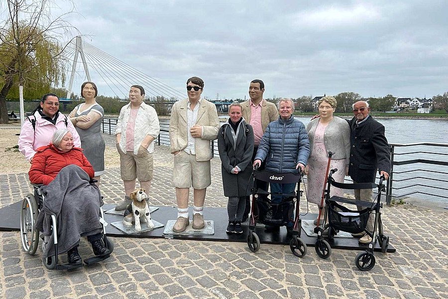 Gruppe von Menschen, darunter zwei mit Rollstuhl, zwei mit Rollator, und mehrere stehende Personen auf gepflastertem Platz an einem Flussufer mit einer Hängebrücke im Hintergrund.