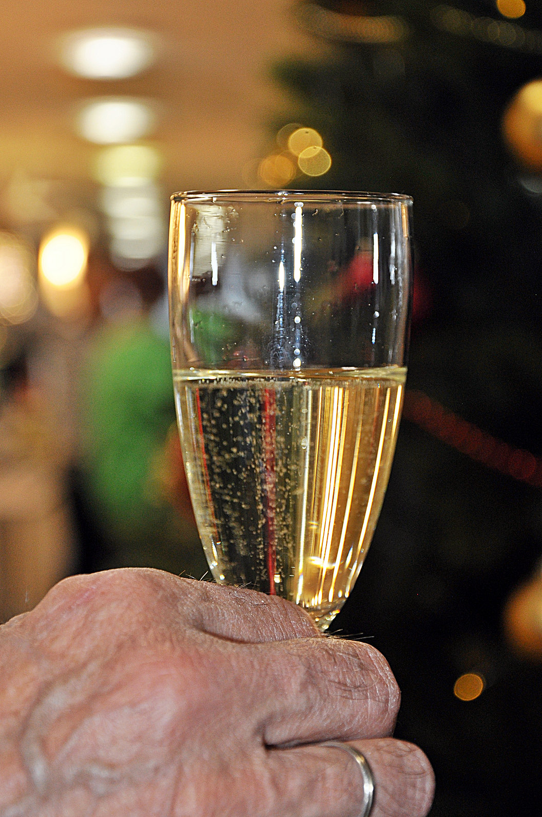 Hand mit Ehering hält ein Glas Sekt vor unscharfem Hintergrund mit Lichtern.