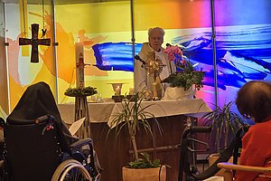 Priester steht hinter Altar mit Monstranz und Kerzen, davor sitzen Menschen, einer im Rollstuhl, in einem Raum mit farbiger Wandgestaltung