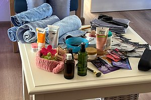 Auf einem weißen Tisch liegen mehrere Kosmetikprodukte, ein rosa Stirnband, ein blauer Schüssel, zusammengelegte Handtücher und ein Kamm