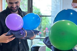 Person hält mehrere bunte Luftballons in einem hellen Raum mit großen Fenstern