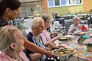 Mehrere ältere Frauen sitzen an einem Gartentisch und eine Frau reicht eine Platte mit belegten Broten.