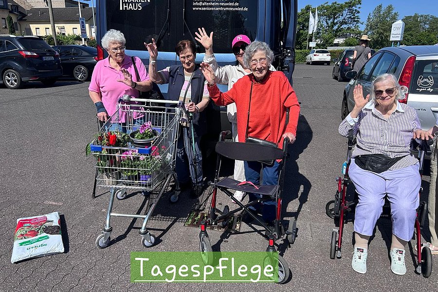 Vier ältere Menschen mit Rollatoren und Rollstuhl stehen auf einem Parkplatz und winken in die Kamera
