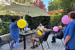 Mehrere ältere Menschen sitzen und stehen im Garten unter einem Sonnenschirm und spielen mit bunten Luftballons