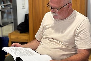 Älterer Mann sitzt an einem Tisch und liest in einem aufgeschlagenen Buch