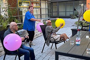 Zwei ältere Männer sitzen auf Stühlen im Außenbereich und spielen mit bunten Luftballons, eine Frau in blauer Kleidung steht daneben.