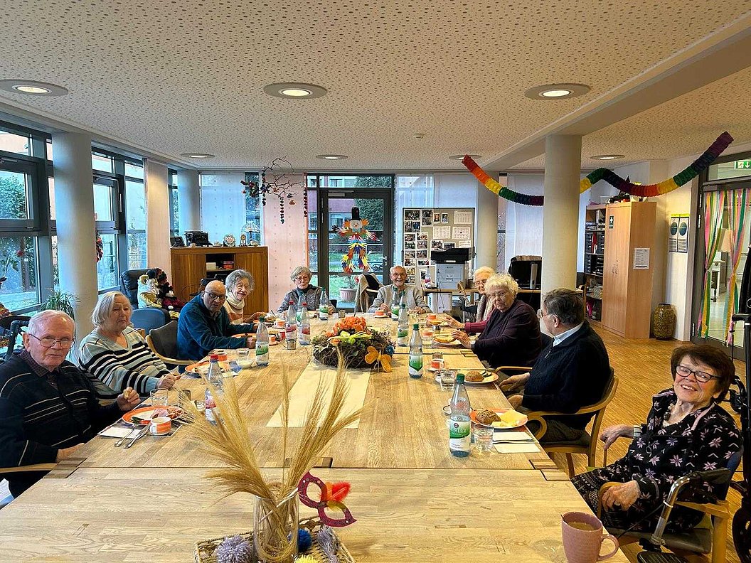 Mehrere Personen sitzen an einem langen Holztisch in einem hellen Raum mit großen Fenstern und dekorativen Elementen an der Decke und Wand.