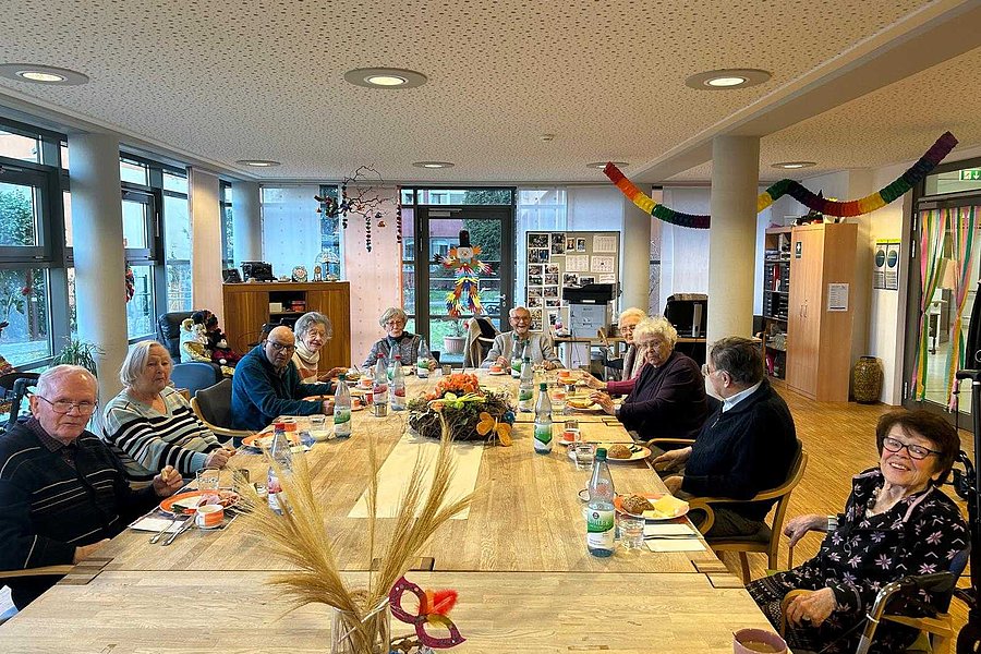 Mehrere Personen sitzen an einem langen Holztisch in einem hellen Raum mit großen Fenstern und dekorativen Elementen an der Decke und Wand.