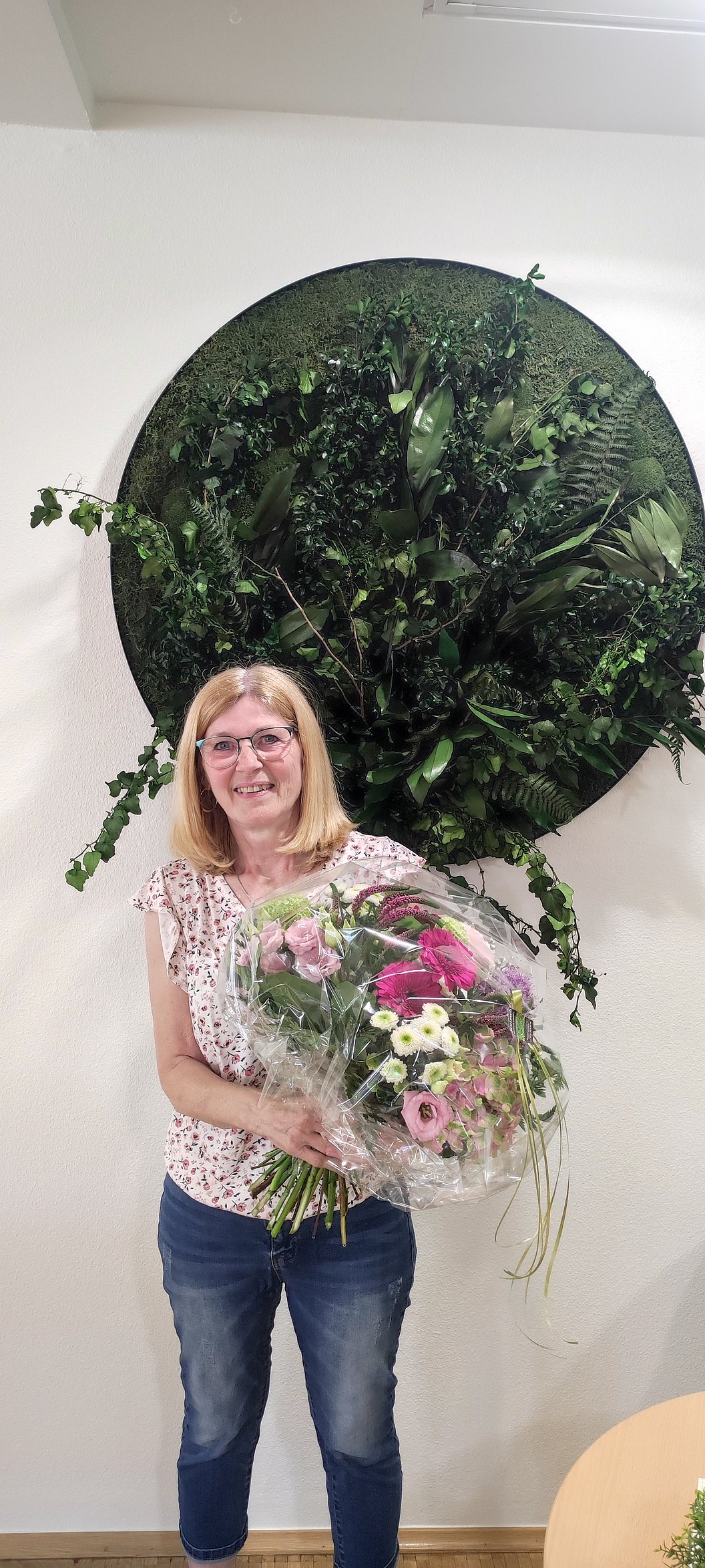 Frau steht vor einer Wand mit rundem, grünem Pflanzenarrangement und hält einen großen Blumenstrauß mit verschiedenen Blüten.