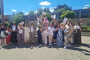 Gruppe von Menschen steht im Freien vor einem Gebäude und hält Papiere in die Luft