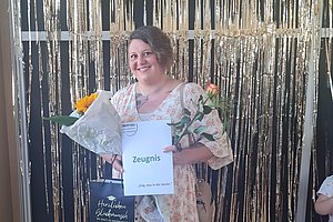 Frau in floralem Kleid hält Blumenstrauß und Zeugnis vor silbernem Lamettavorhang mit weißen und goldenen Luftballons