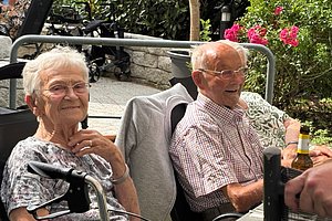 Zwei ältere Personen sitzen draußen an einem Tisch, eine Person hält eine Bierflasche, im Hintergrund Blumen und Gartenmöbel.