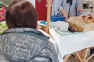 Seniorin hinter einem Rollstuhl, am Tisch mit Lebensmitteln und einer Verkäuferin in einem Marktsetting.