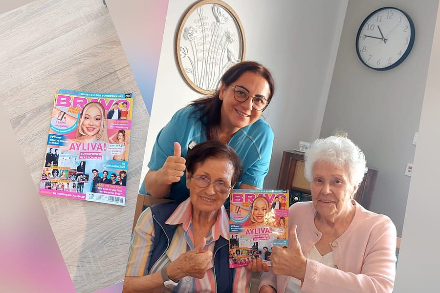 Drei Frauen sitzen an einem Tisch, zwei ältere Frauen halten jeweils eine Zeitschrift, eine jüngere Frau steht hinter ihnen und lächelt